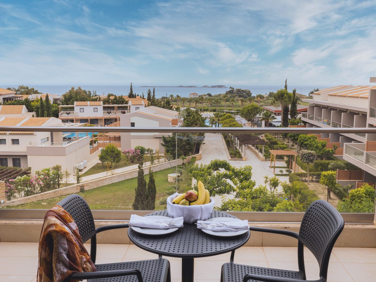 A furnished room balcony with a table and fruit bowl, looking out over the resort's terraced gardens and pool to the Ionian sea and a small islet.