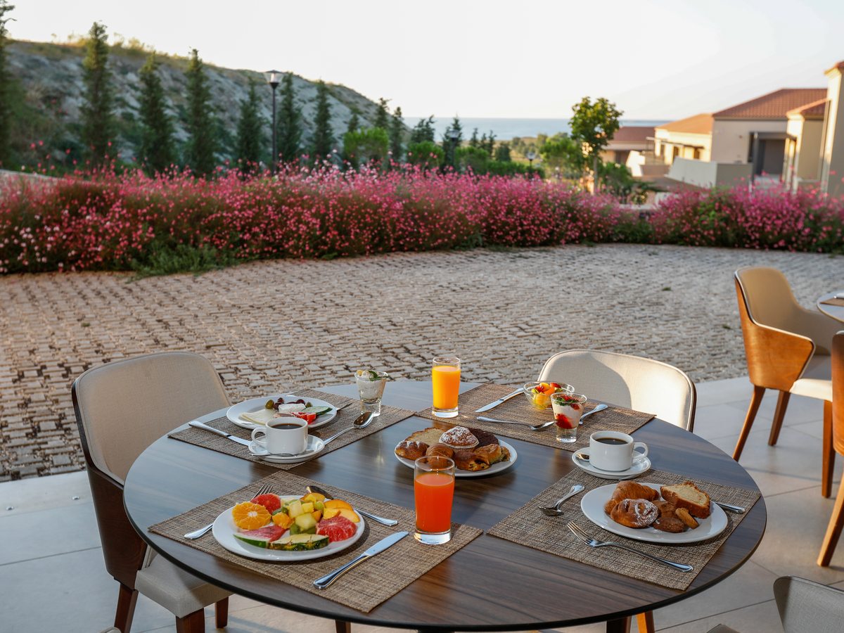 An outdoor breakfast table set with pastries, fruit, coffee and juices, with the resort's flowering gardens and countryside beyond.