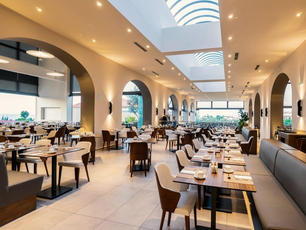 A bright modern dining room with arched windows, a central skylight, wooden tables, pale chairs and grey banquettes.