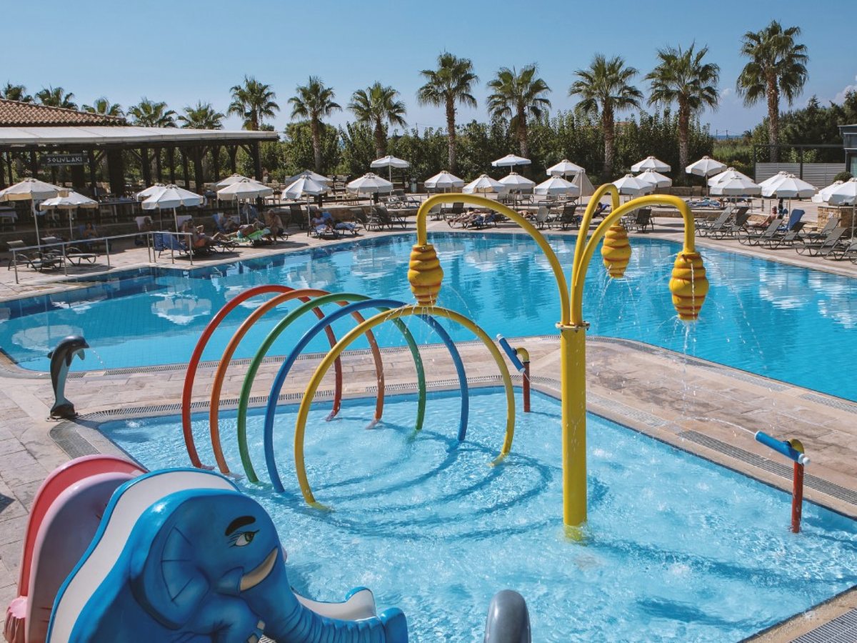 A shallow children's splash pool with colourful hoops, water-spray features and an elephant slide, beside a larger pool.