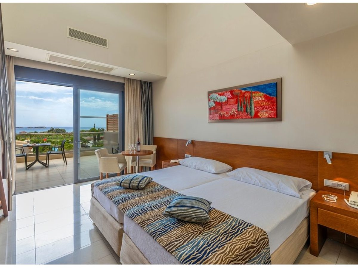 A high-ceilinged room with a wooden internal staircase to one side, twin beds and a sea-view balcony with a small table.