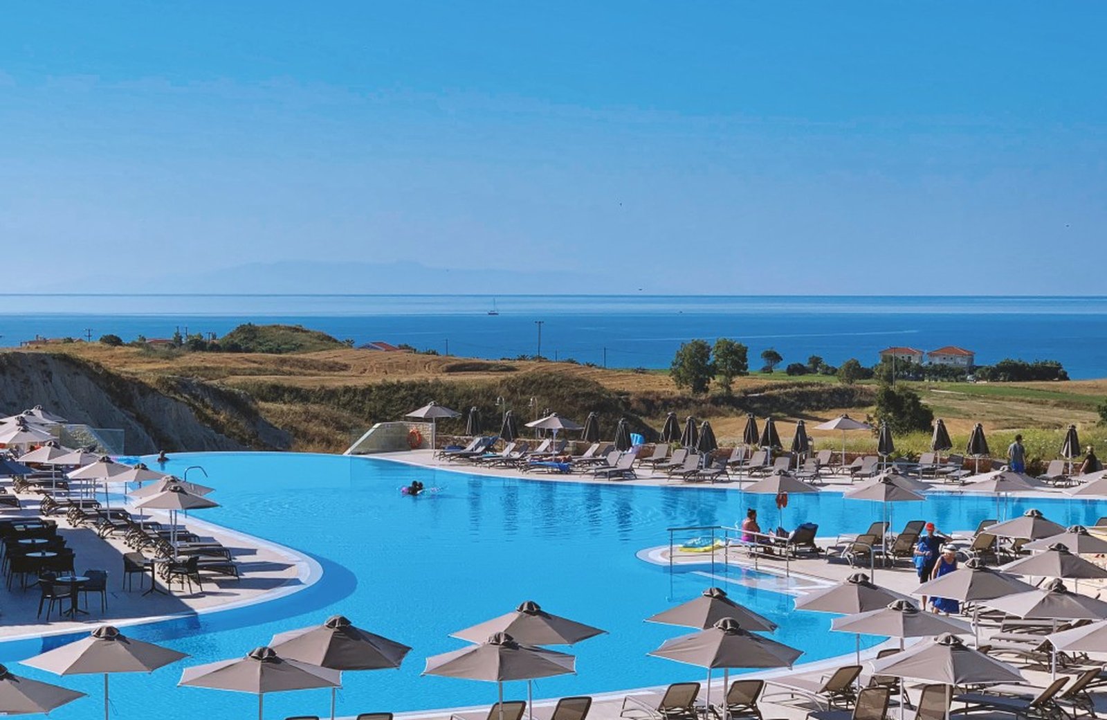 A large curved resort pool ringed with sun loungers and parasols, looking out over open fields to the Ionian sea on the Paliki Peninsula in Kefalonia.
