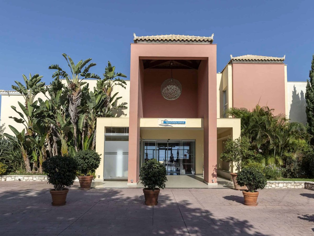 A tall peach entrance portico with glass doors, flanked by potted palms and planting.