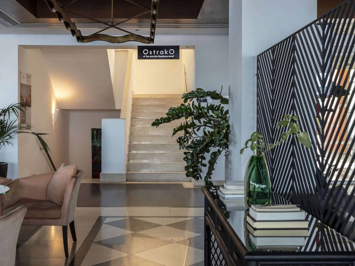A restaurant entrance with an Ostrako sign above a short flight of stairs, an armchair and potted plants in the foyer.
