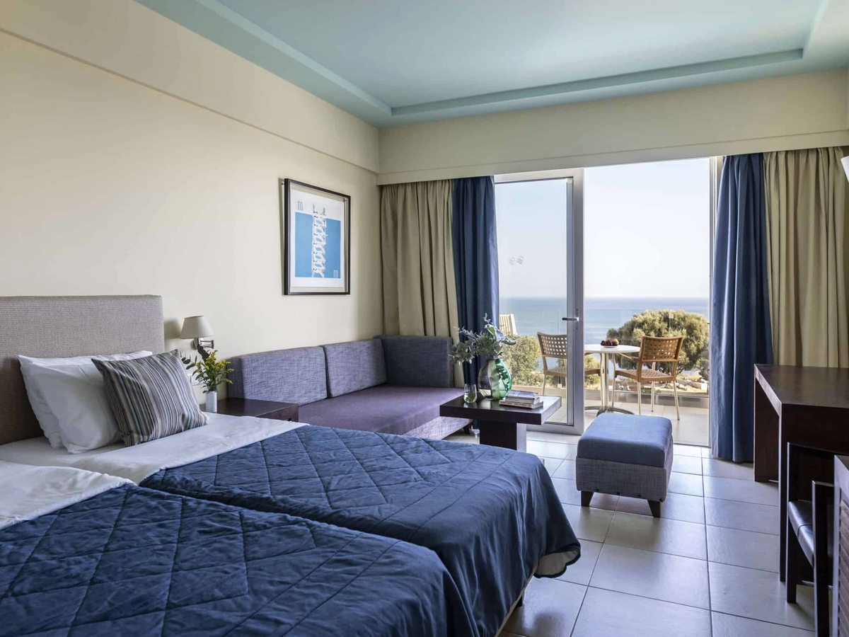 A room with two single beds under blue covers, a sofa bed by the window and doors onto a private sea-view balcony.