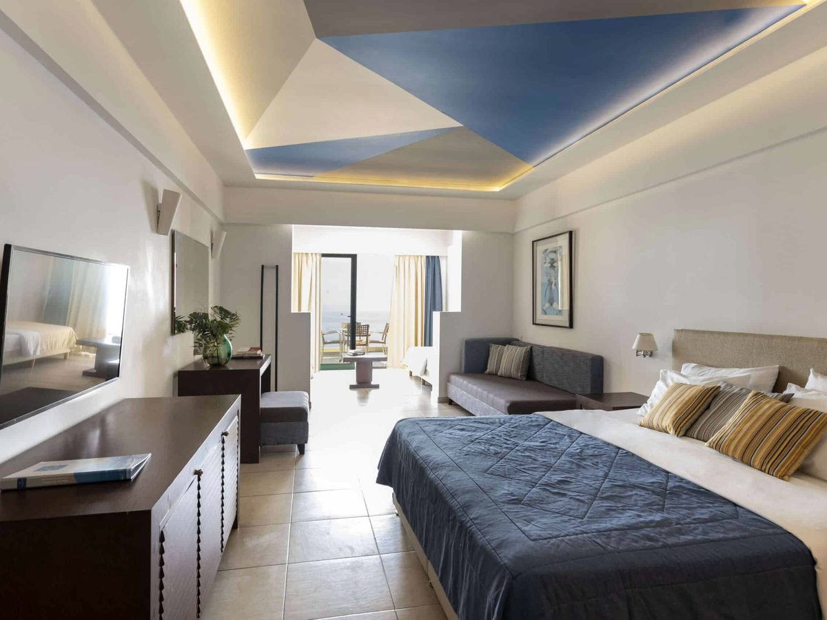 A large room with a double bed, a sofa and seating area, a long desk and sliding doors to a sea-view balcony.