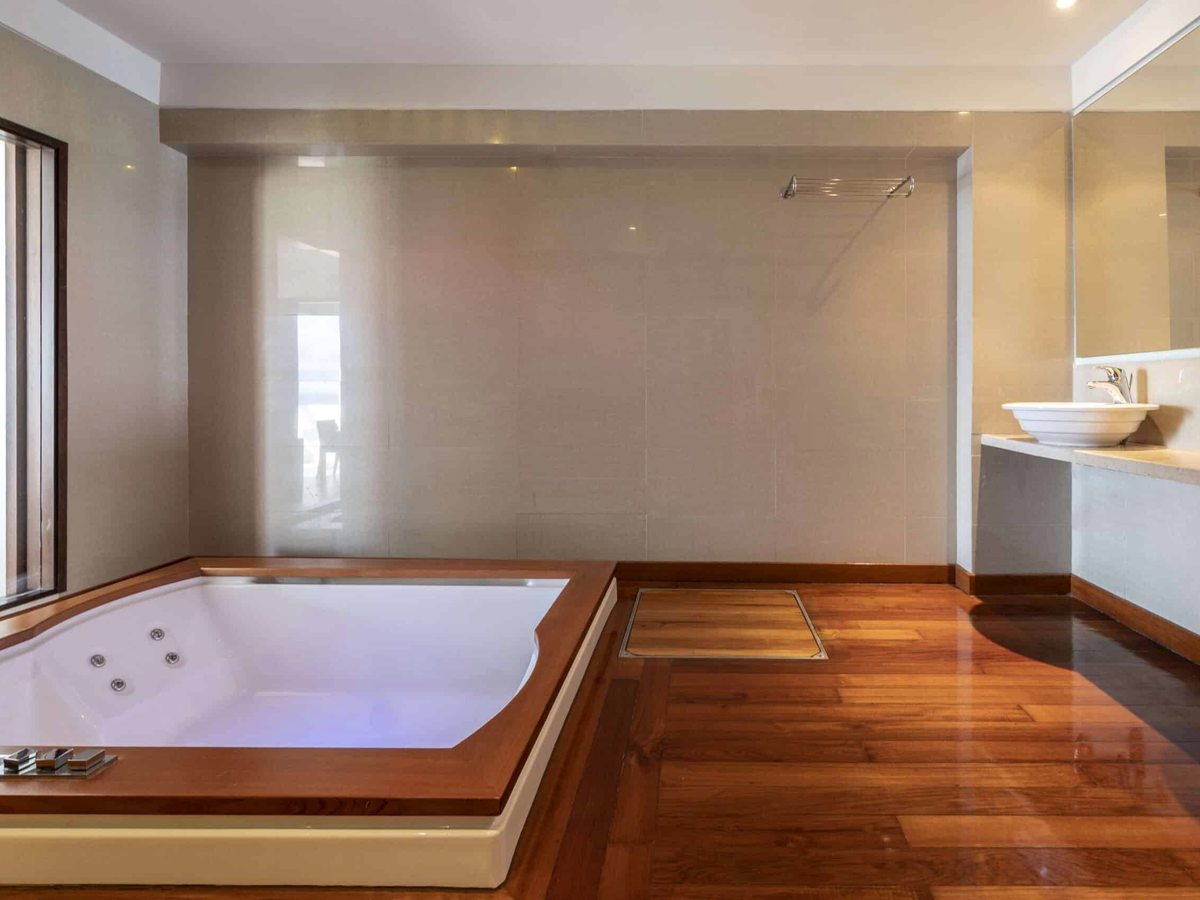 A large sunken whirlpool bath with a wooden surround against a tiled wall, beside a vanity basin, with a window to the side.