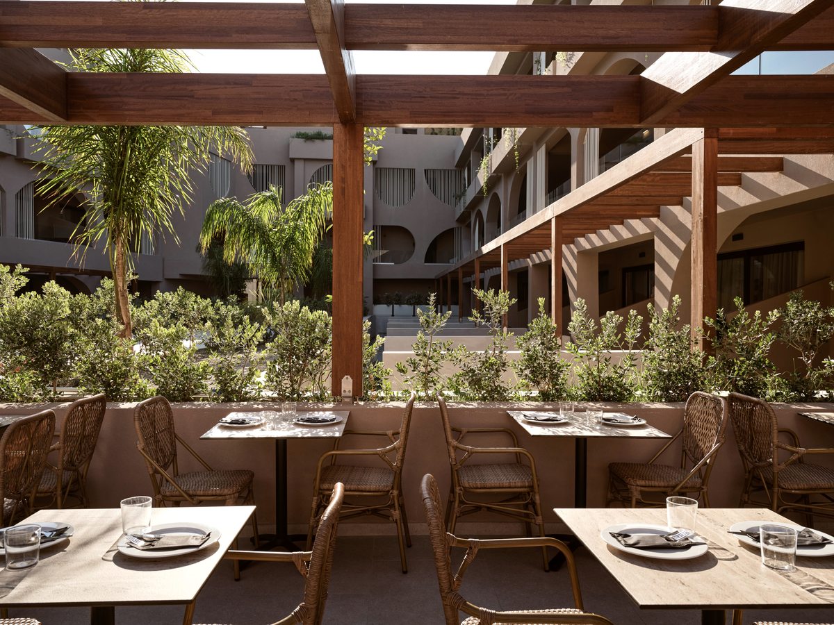 An outdoor restaurant terrace set for a meal, with rattan chairs and laid tables under a timber pergola beside planted borders, the room blocks beyond.