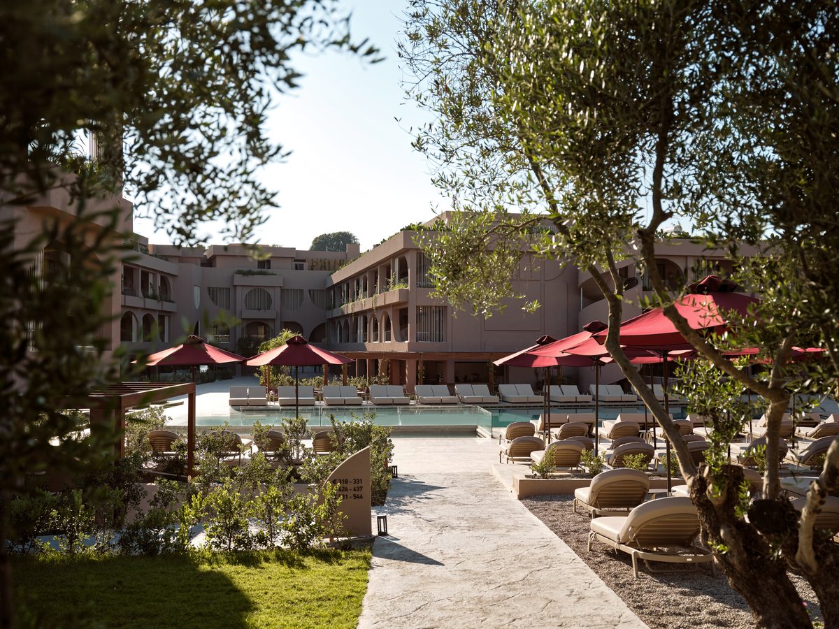 A pool set among olive trees and lawns, with red parasols and rows of sun loungers, the peach room blocks behind.