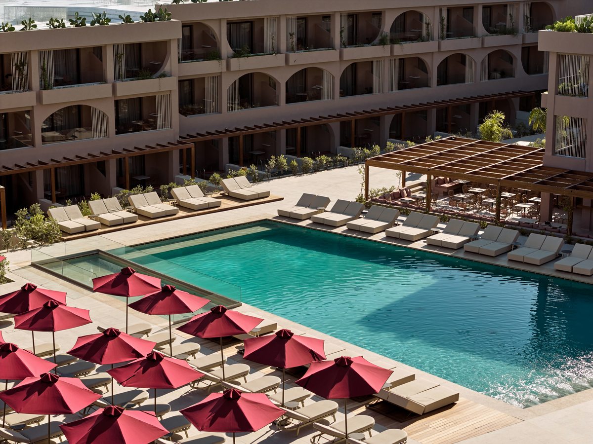 A large rectangular pool with rows of sun loungers and red parasols along the deck, a timber pergola to one side and the multi-storey room blocks behind.