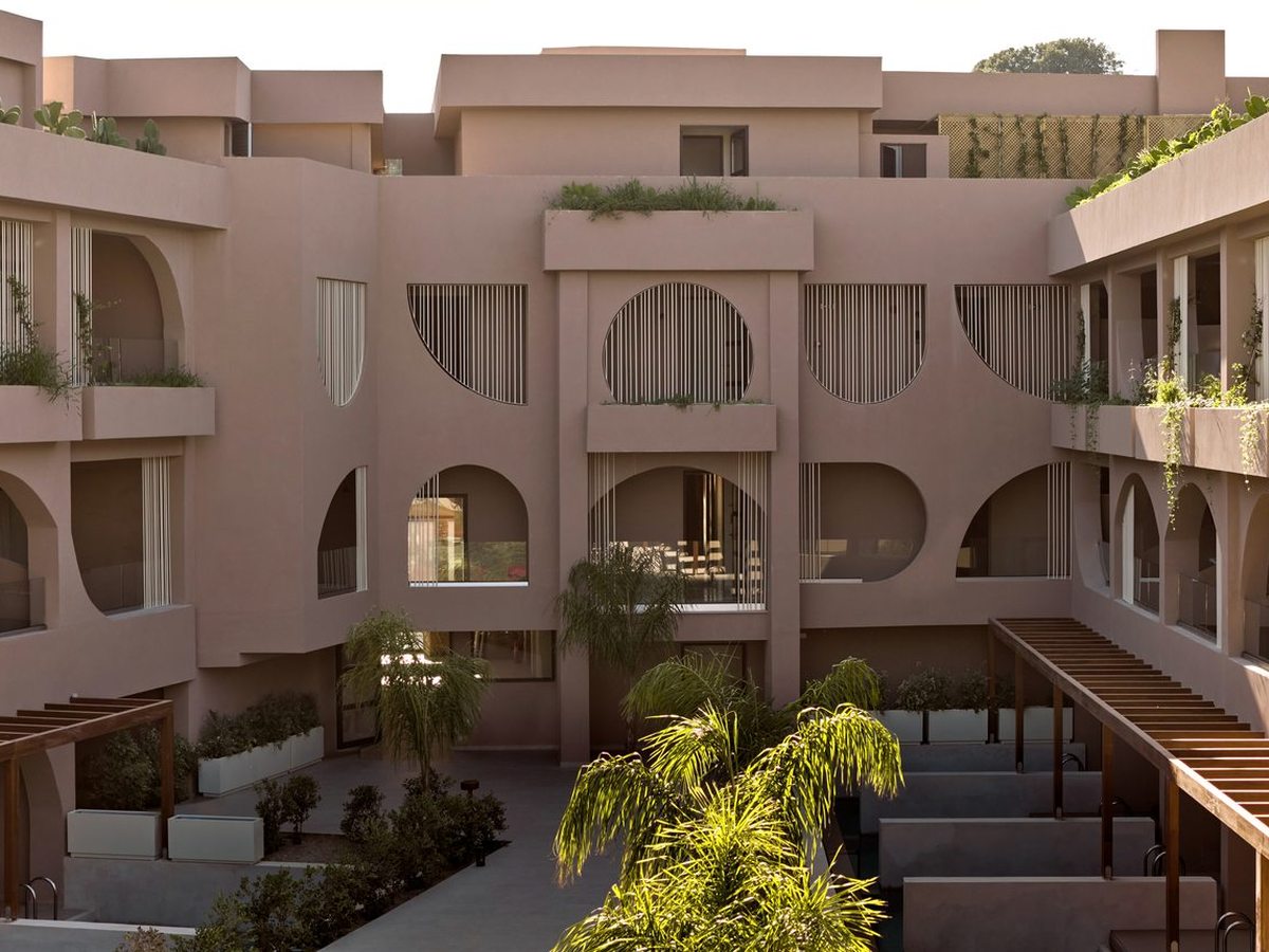 A raised walkway looking across an internal courtyard of arched-balcony room blocks, with palms and planted borders under a clear sky.