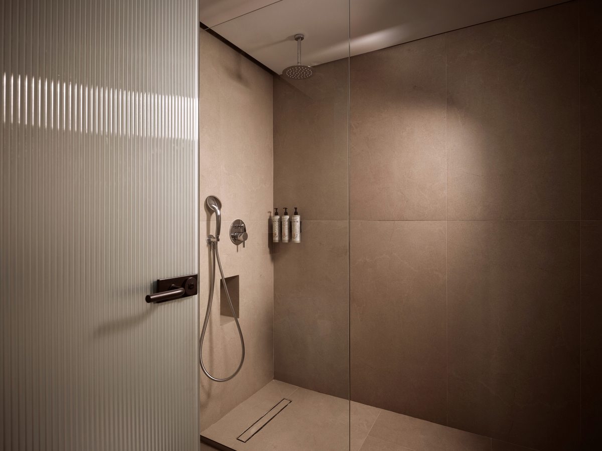 A tiled walk-in shower with a rainfall and handheld head behind a fluted glass screen, a linear floor drain and wall-mounted dispensers.