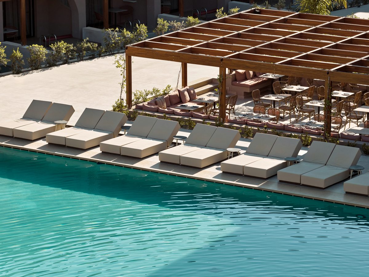 Rows of double sun loungers along the edge of a pool, beside a large timber pergola sheltering a lounge and dining area.