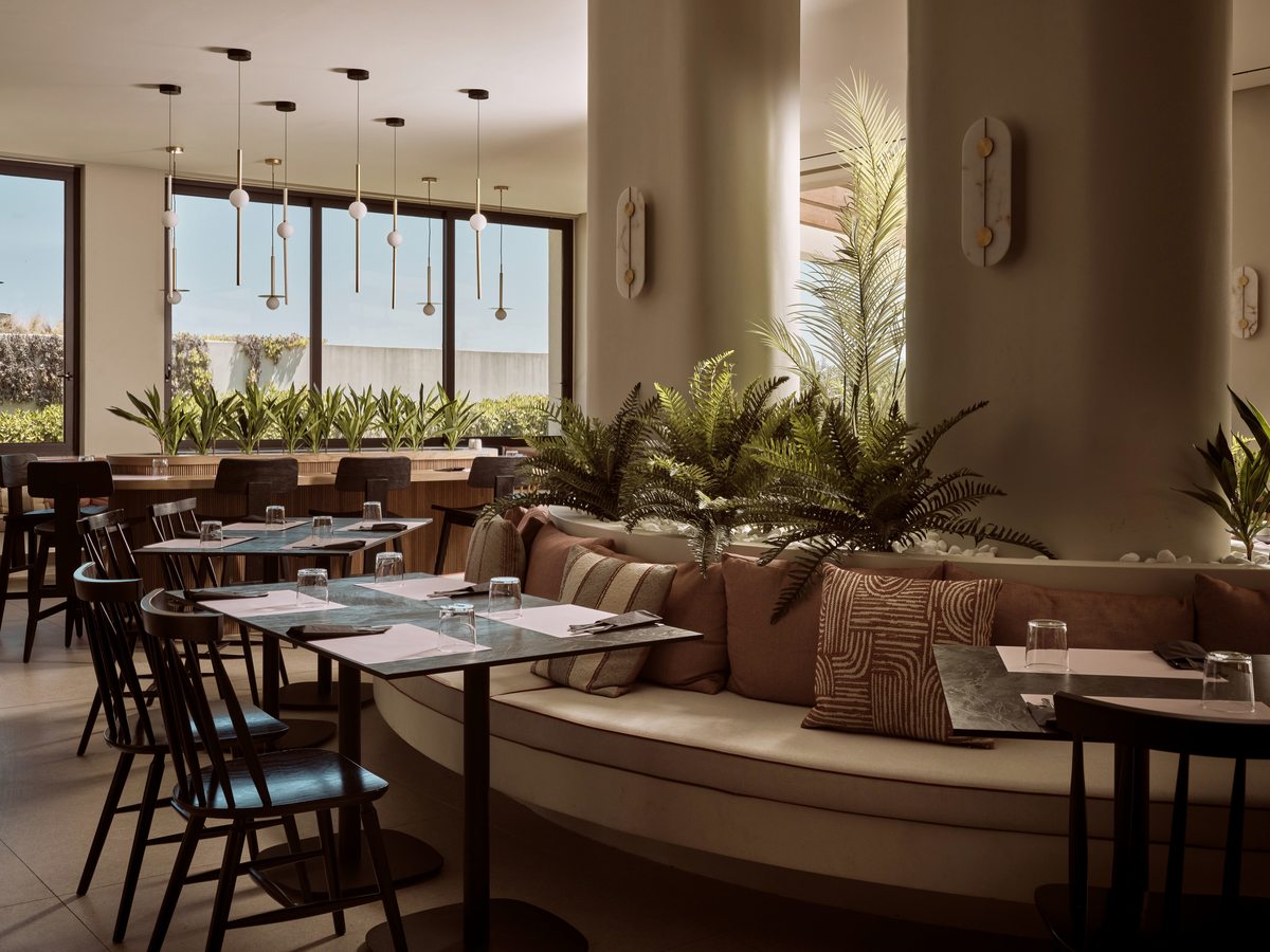 An indoor restaurant with laid tables, curved banquette seating and black dining chairs under pendant lights, with tall windows and planted dividers.