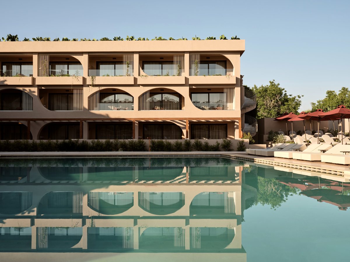 A peach-rendered three-storey apartment block with arched balconies and rooftop planting, its rows of rooms reflected in the pool below and sun loungers along the near deck.