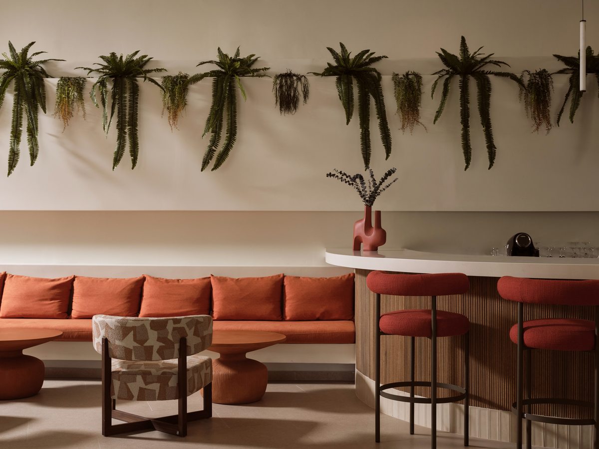A lounge bar with a rust-orange banquette, red stools at a curved white counter and patterned armchairs, hanging plants along the wall.