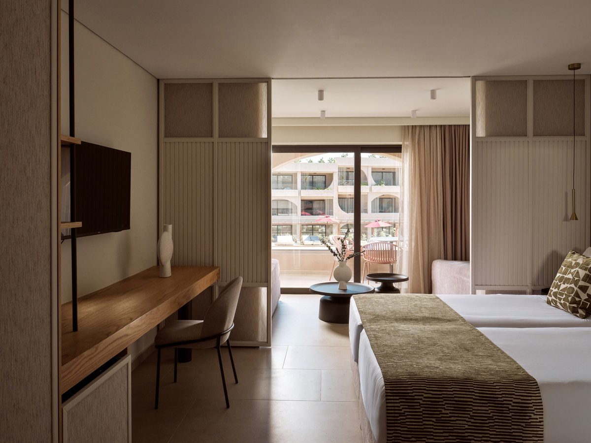 A bedroom with a wall-mounted TV over a timber desk and sliding doors to a balcony that looks onto the pool and the arched room blocks.