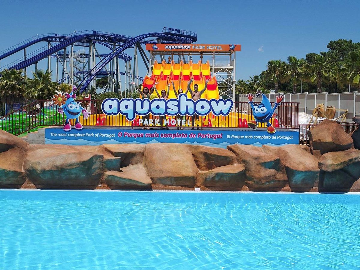 A large pool with rock-effect edging in front of the aquashow Park Hotel sign, with yellow drop slides and a roller-coaster track behind.