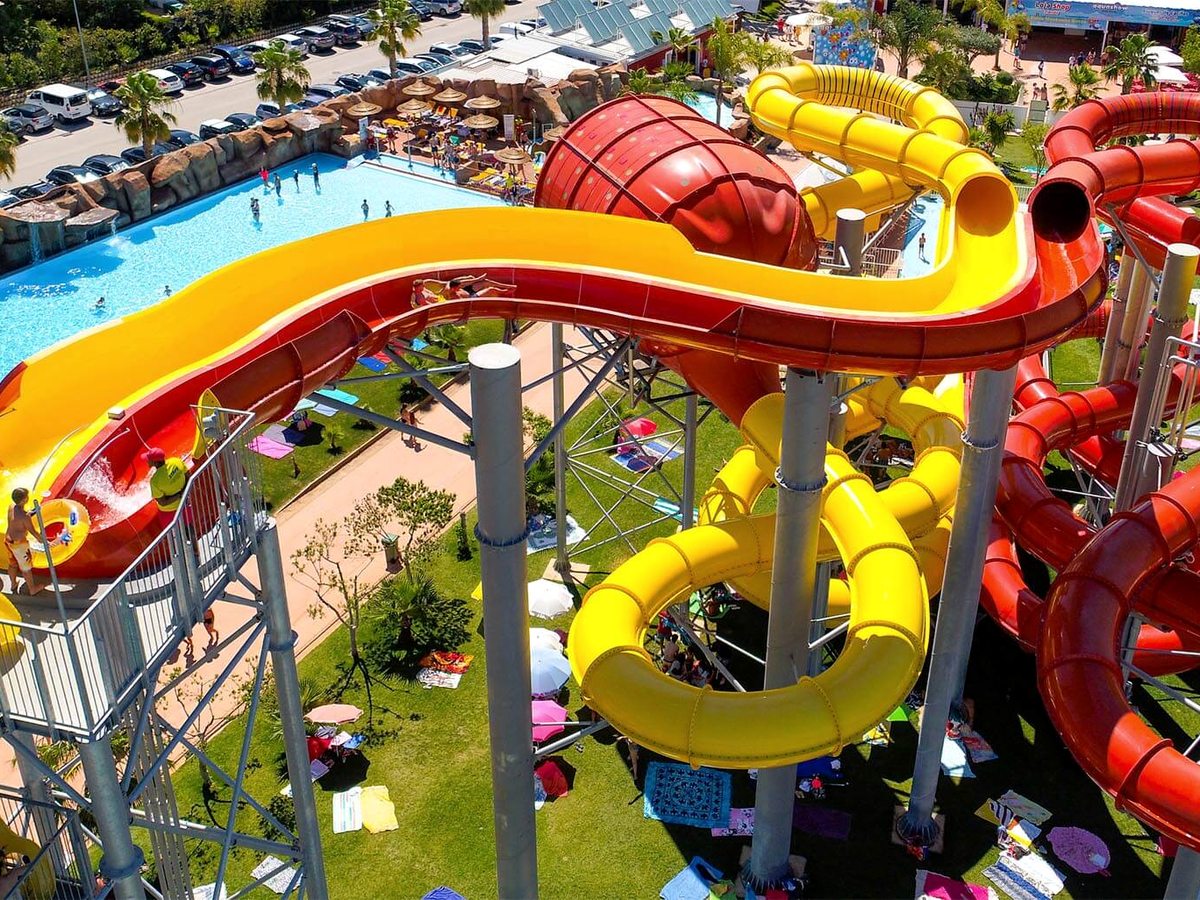 Yellow and red enclosed tube slides spiralling down beside an open body slide among palm trees.
