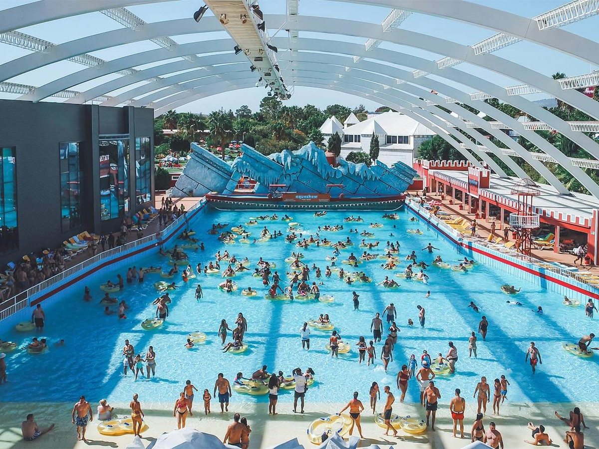 A large covered wave pool busy with people on yellow tube rings under a curved translucent canopy.