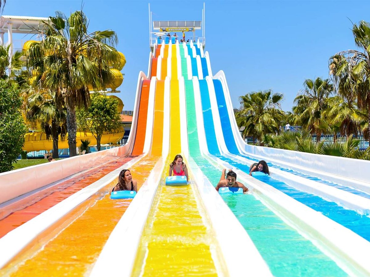 Several children lying on mats racing down a rainbow multi-lane slide.