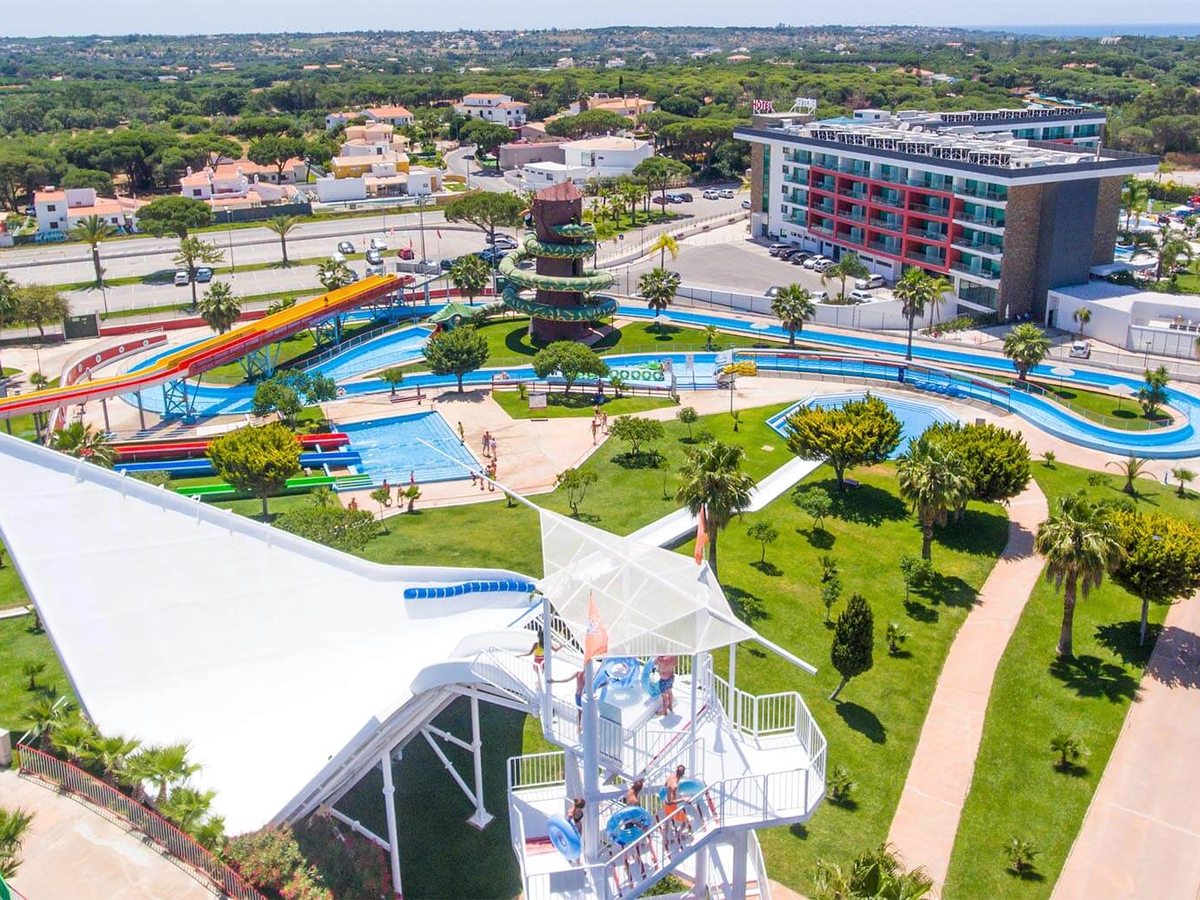 Aerial view of the multi-storey Aquashow Park Hotel beside its water park, with a lazy river, slide tower and pools among lawns and Quarteira beyond.