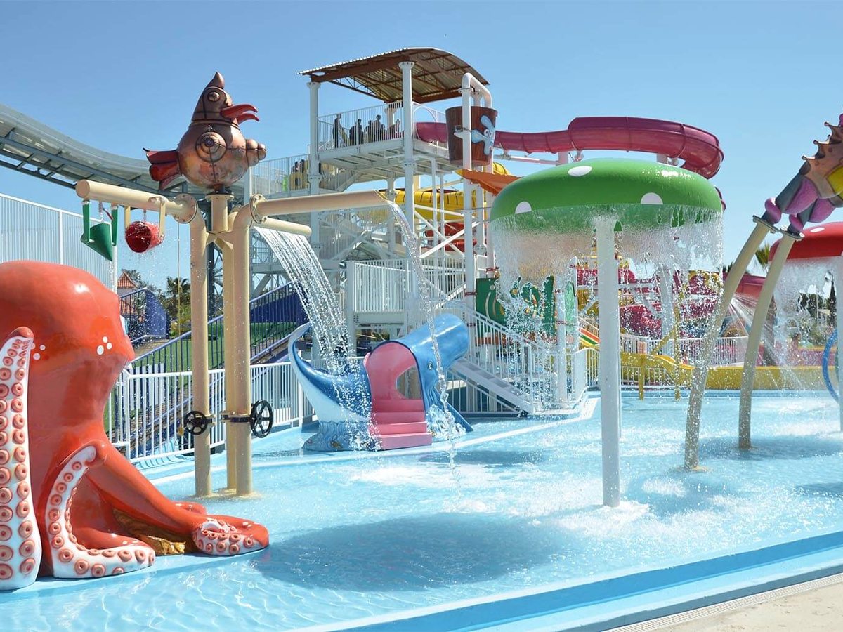 A shallow water-play area with an octopus feature, mushroom sprayers and small slides behind white railings.