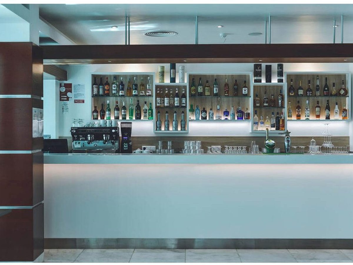 A hotel lobby bar with a white counter, backlit shelves of spirit bottles and a coffee machine.