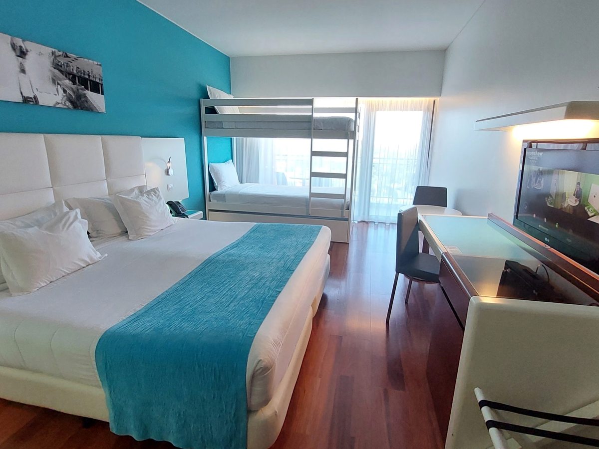 A Twin Plus room with two single beds pushed together under a turquoise runner, a separate metal bunk bed against a turquoise wall, a desk, chair and balcony door.