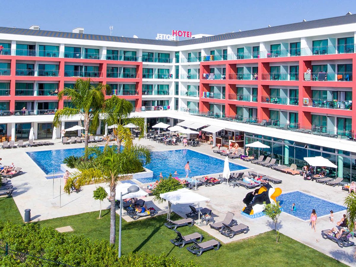The hotel's outdoor pool in a courtyard below the red-and-turquoise building, with sun loungers, parasols and palm trees.