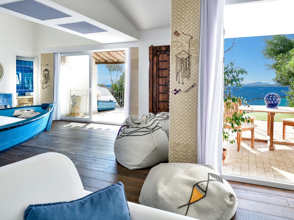 An airy open-plan suite where a blue-painted wooden fishing boat serves as a bench, with bean-bag seats, a carved door and sliding doors to a sea-view terrace.