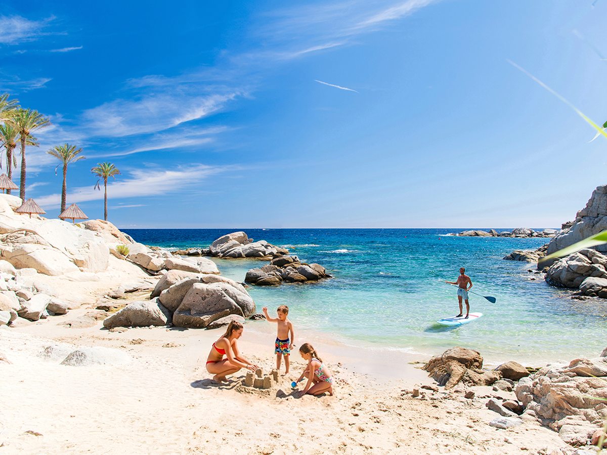 People on white sand beside a rocky cove, with a paddleboarder on calm turquoise water and palms behind.