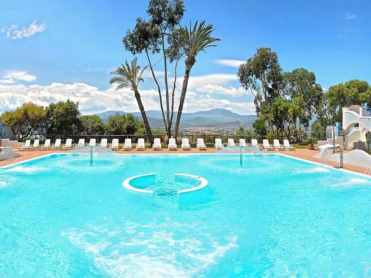 A large rectangular pool ringed with white loungers on a terrace, with tall palms and a mountain skyline beyond.