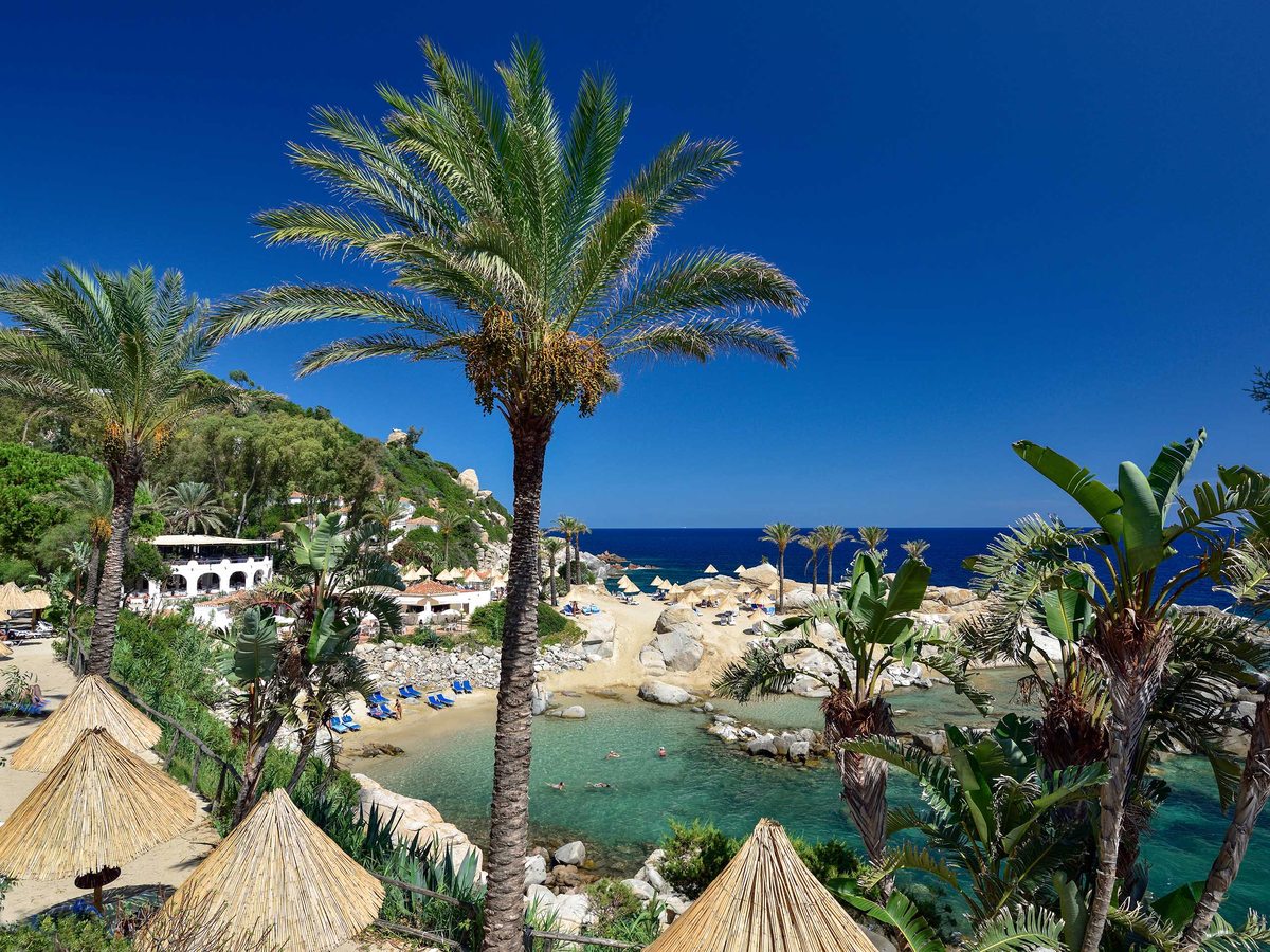 A sheltered turquoise cove ringed by pale boulders and date palms, with swimmers in the water and thatched umbrellas on the sand.