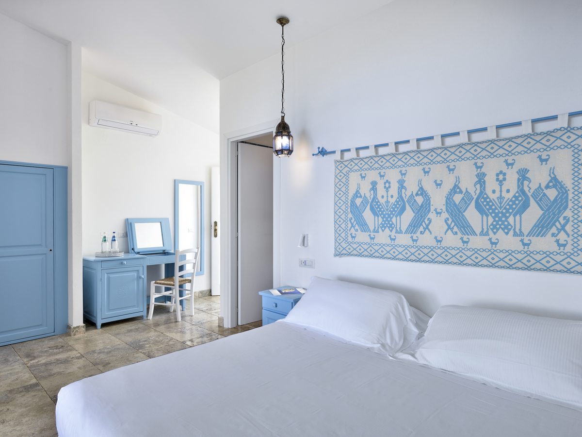 A double bedroom with a blue-and-white folk-motif textile panel above the bed, a blue-painted dresser and wardrobe and a travertine floor.