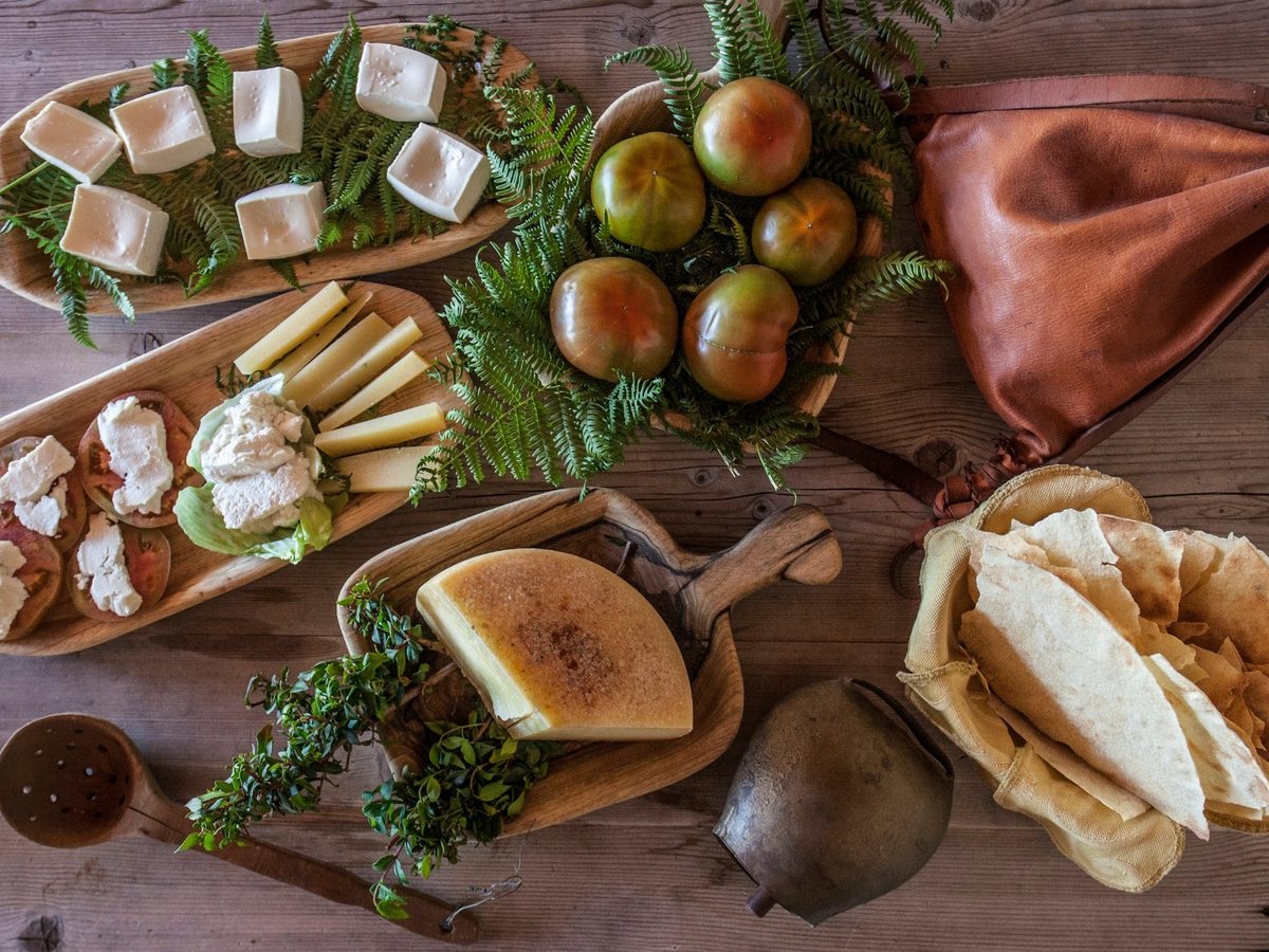 A wooden board of Sardinian cheeses, tomatoes, herbs and thin flatbread laid out on a table.