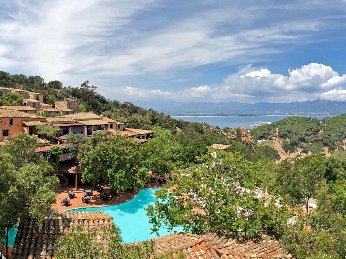 Elevated view of terracotta and pink resort buildings among green trees on a hillside, with a turquoise bay and mountains beyond.