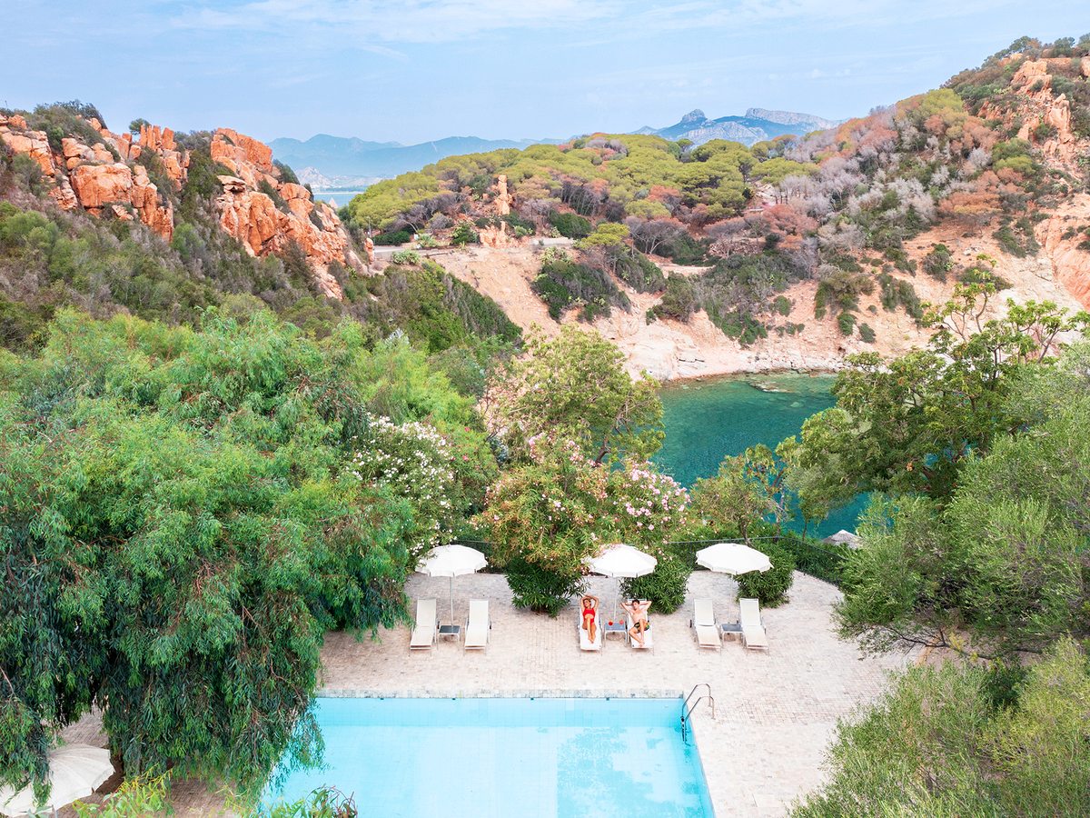 A small pool with loungers and white parasols tucked below wooded red-rock cliffs, with a green cove and mountains beyond.