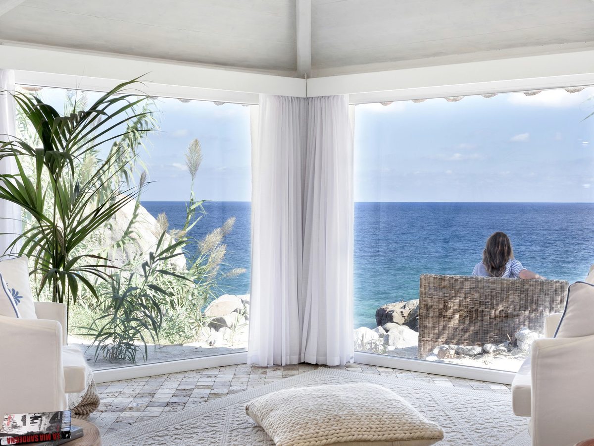 A white-walled suite with two full-height corner windows looking straight out over the sea, cream armchairs and a woven floor cushion.