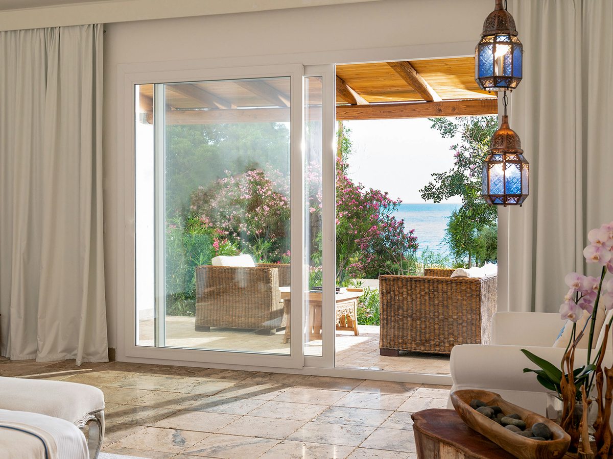 A bright bedroom with sliding glass doors opening onto a furnished sea-view terrace, with a hanging lantern and a pale tiled floor.