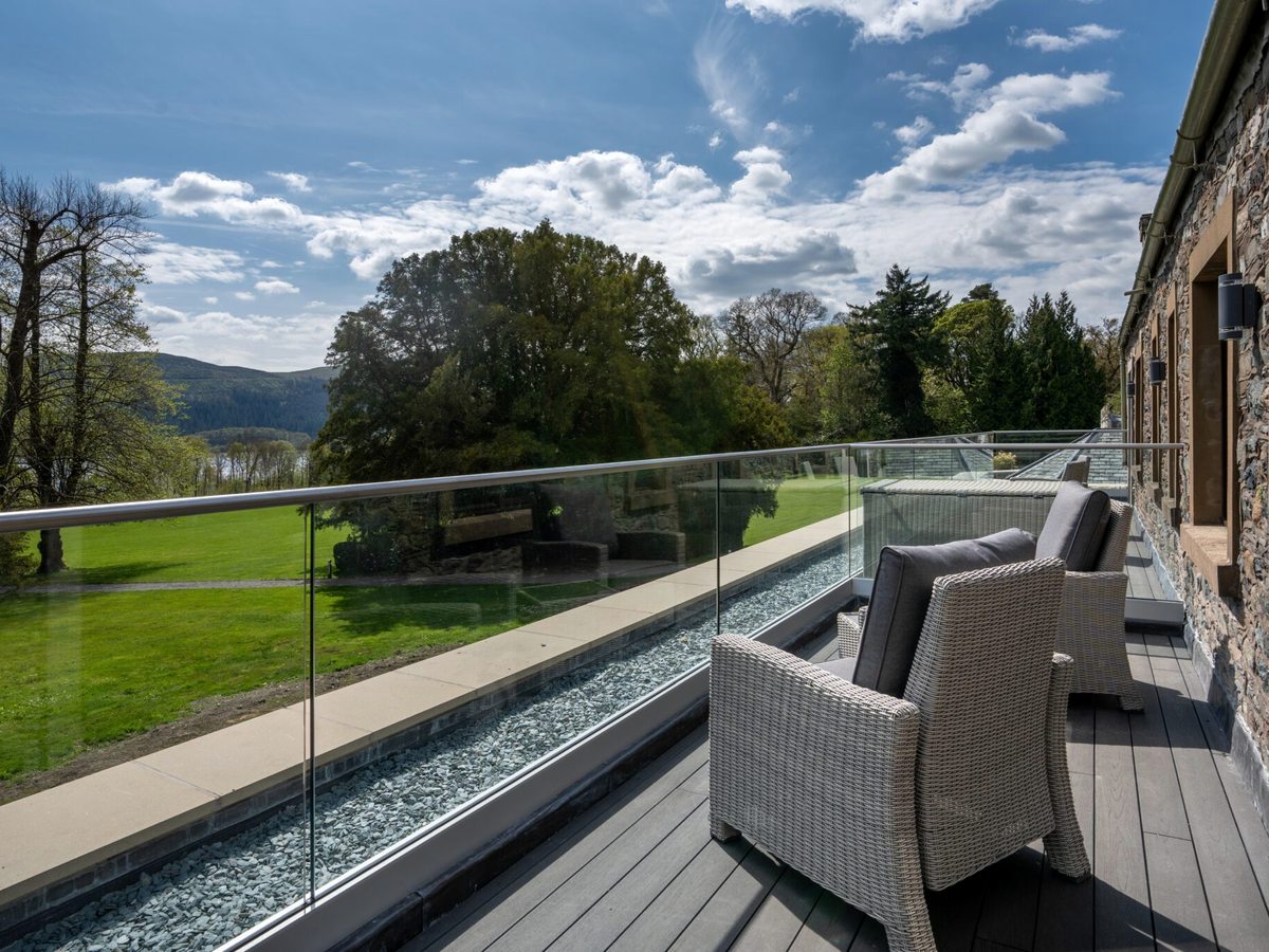 A decked terrace with wicker armchairs and a glass balustrade, looking over lawns to the lake and hills.