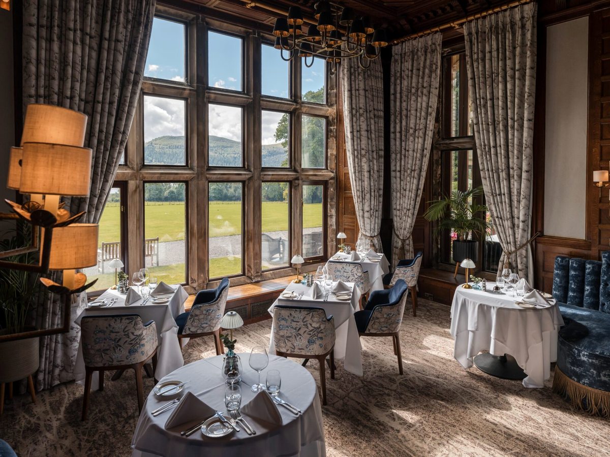 A wood-panelled fine dining room with white-clothed tables and tall windows onto a lawn and fell.