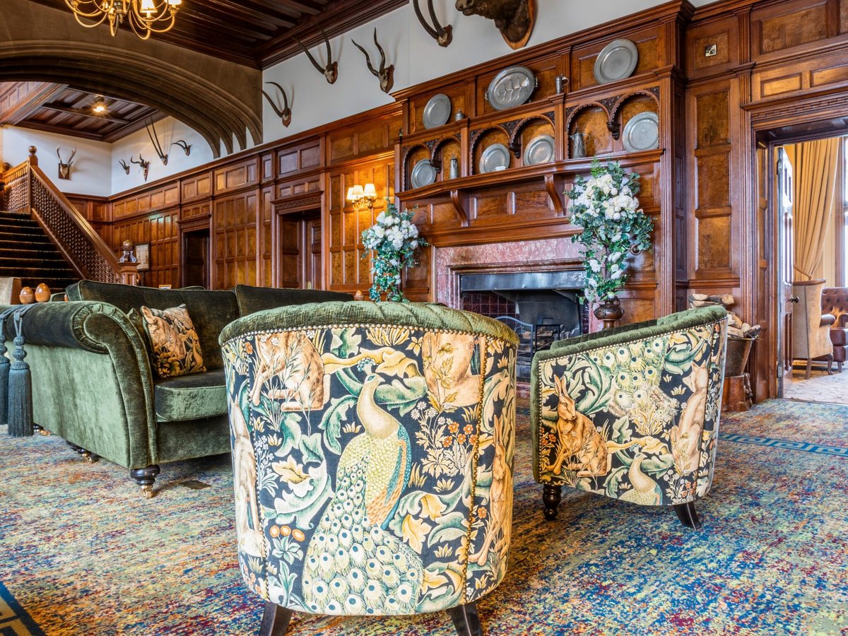A wood-panelled lounge with mounted antlers, a carved stone fireplace, patterned tub chairs and a staircase beyond.