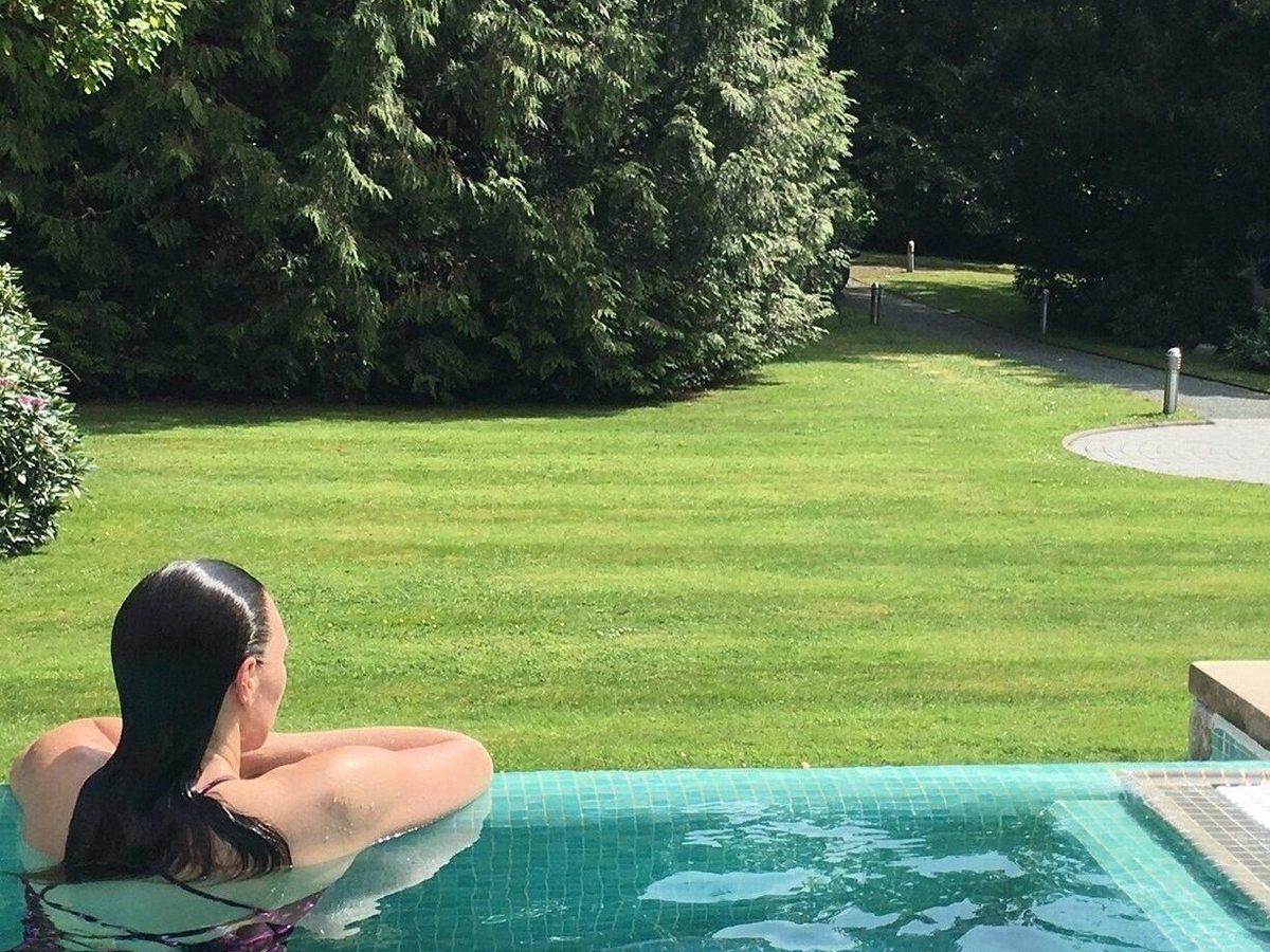 A person resting at the tiled infinity edge of an outdoor pool looking over a lawn and trees.