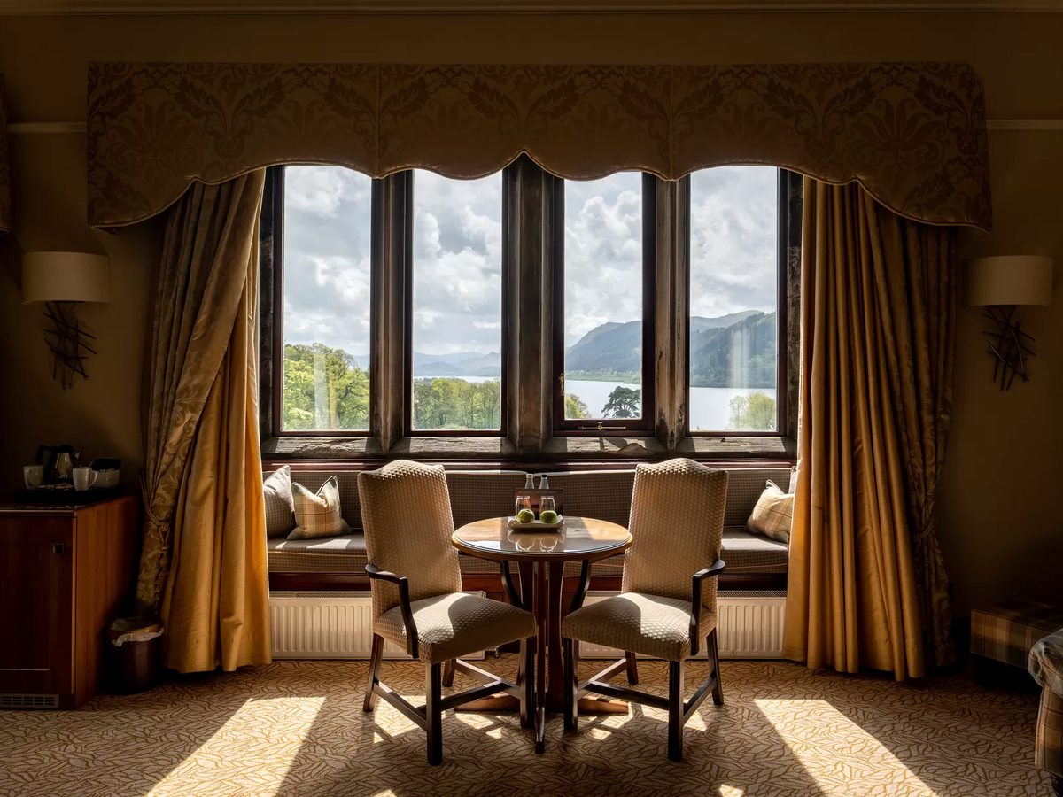 A bay window with a cushioned window seat, a small table and two chairs, the lake and fells beyond.