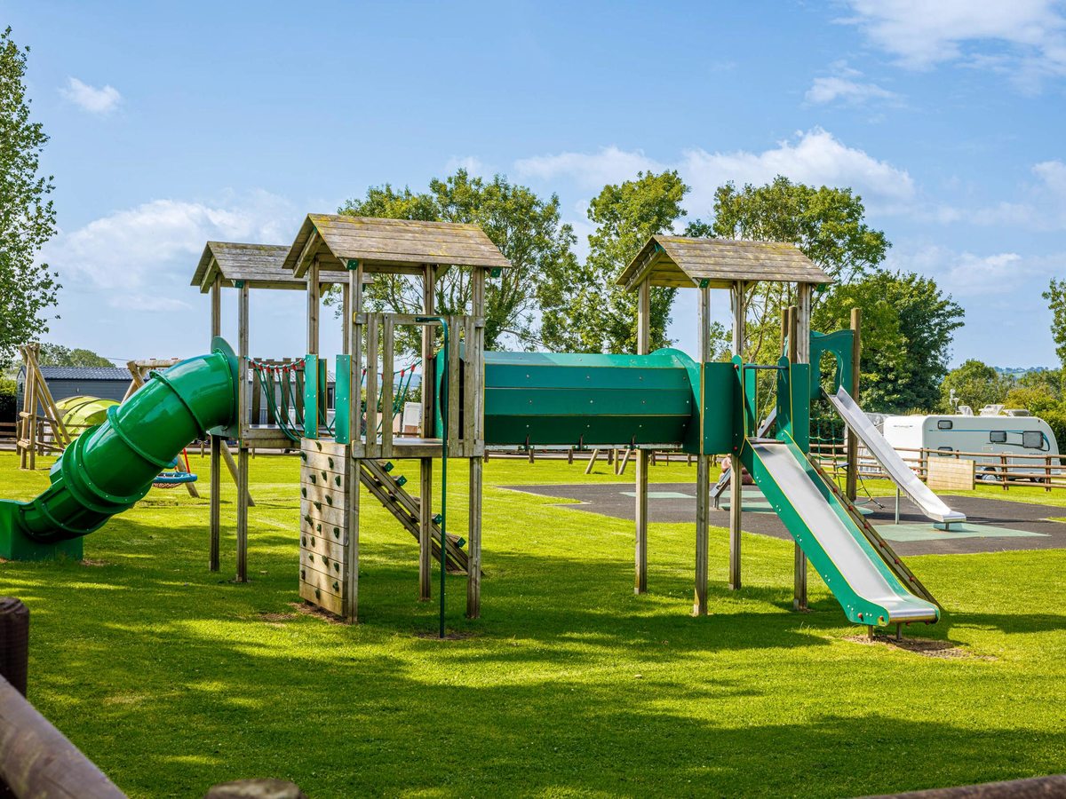 An outdoor play area with wooden climbing towers, a green tube slide and a straight slide on grass, static caravans behind.