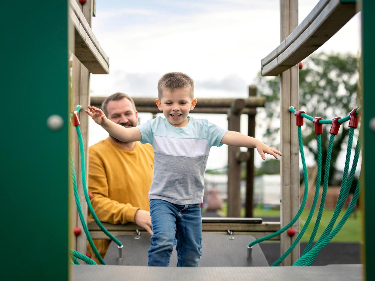 A smiling young boy crossing a rope bridge on a wooden play frame with his father behind him, open grass beyond.