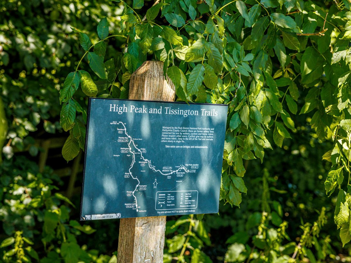 A green trail information board for the High Peak and Tissington Trails on a wooden post beside a leafy hedge.
