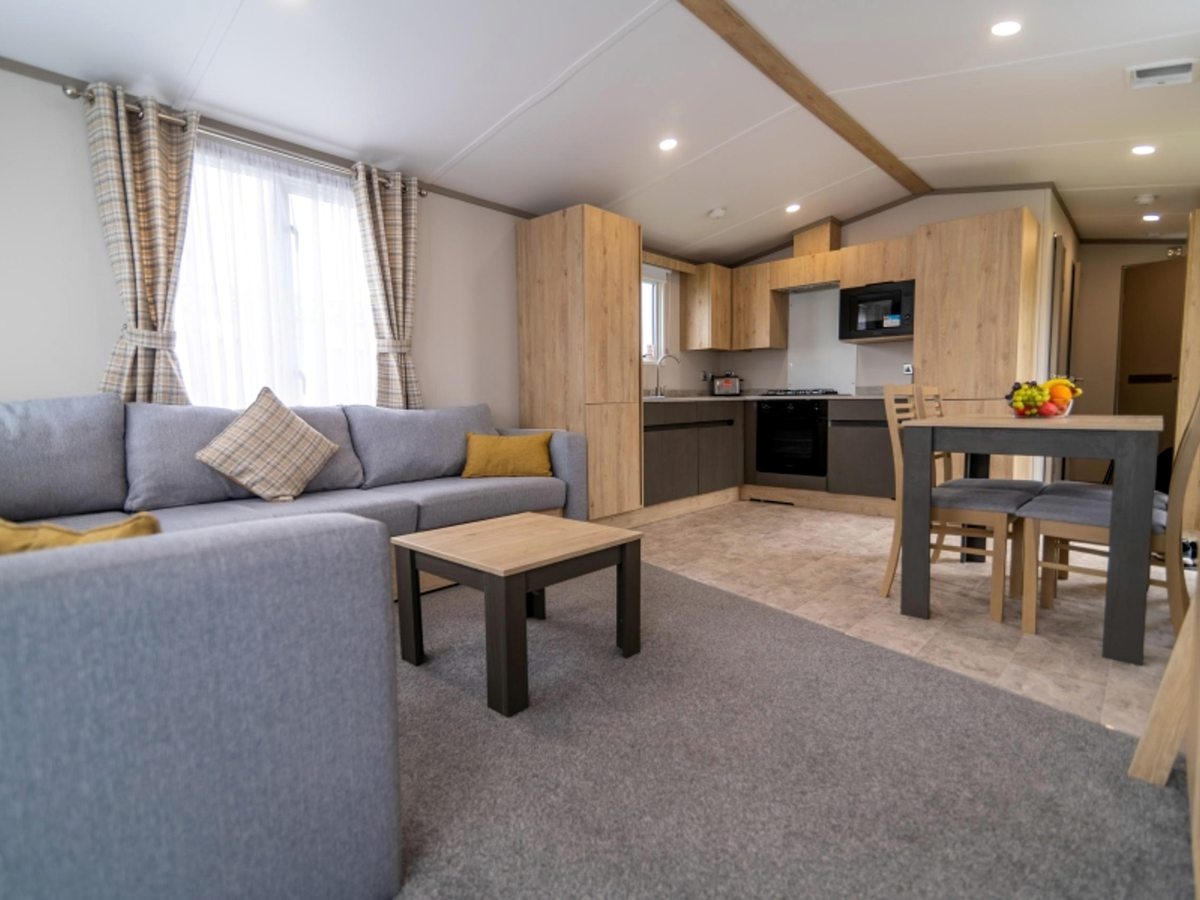 An open-plan static caravan interior with a grey corner sofa, coffee table, dining table and a fitted kitchen with wood cabinets.