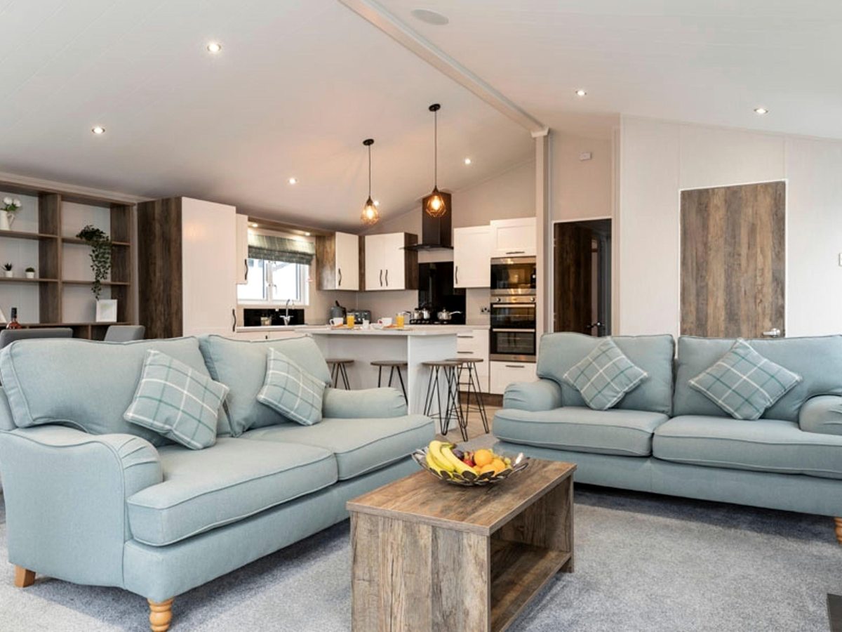 A spacious lodge living area with two duck-egg sofas, a wooden coffee table and a fitted kitchen with a breakfast bar under a vaulted ceiling.
